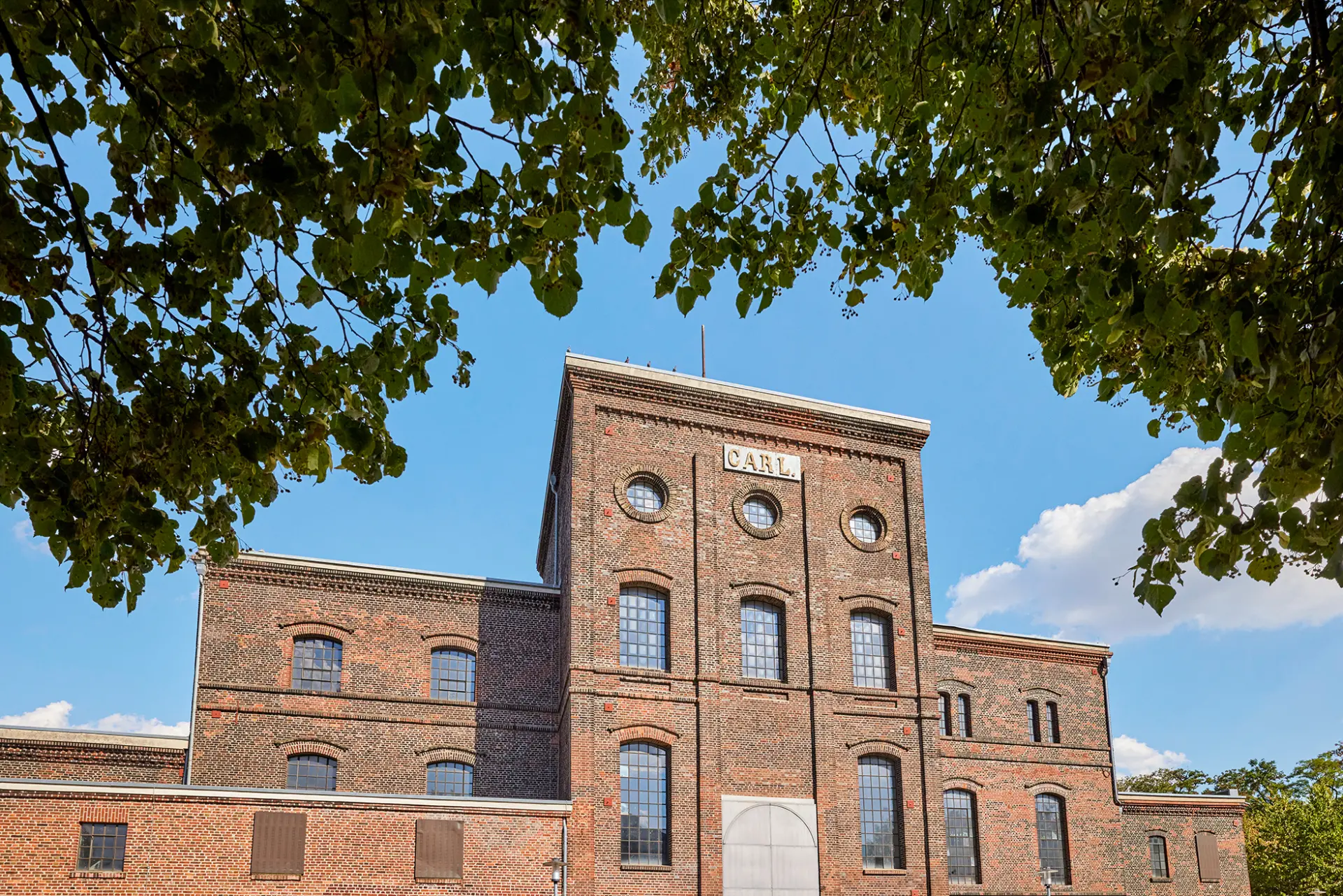 Ehemalige Zechengebäude mit Ziegelarchitektur, blauer Himmel im Hintergrund, eingerahmt von Baumblättern.