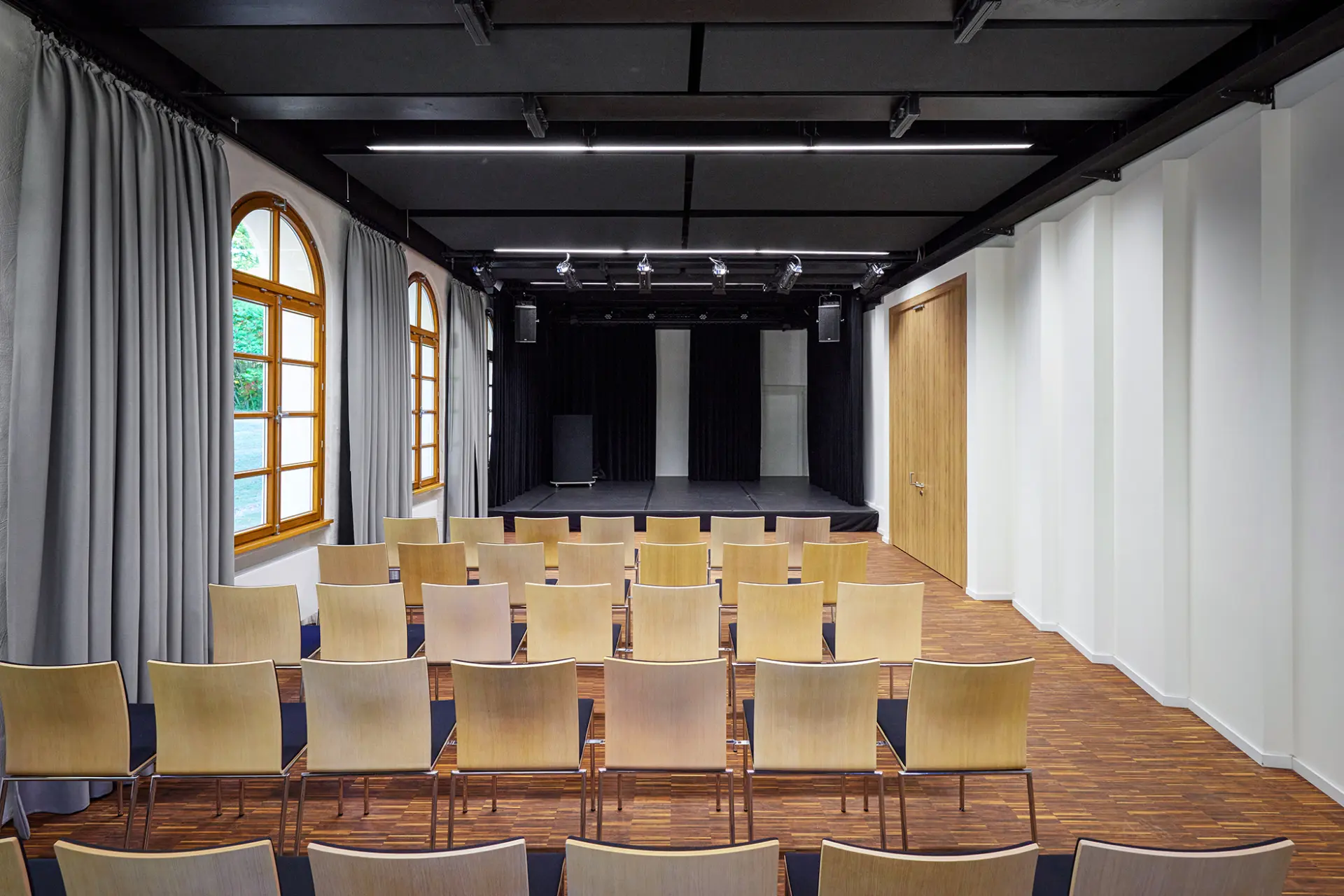 Moderner Veranstaltungssaal mit Holzboden, beige Stühlen und Bühnenbereich inklusive Lautsprechern.