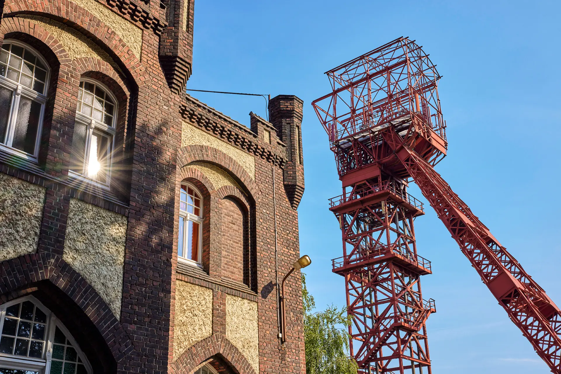```html Historisches Backsteingebäude und roter Bergbau-Förderturm vor strahlend blauem Himmel in Essen. ```
