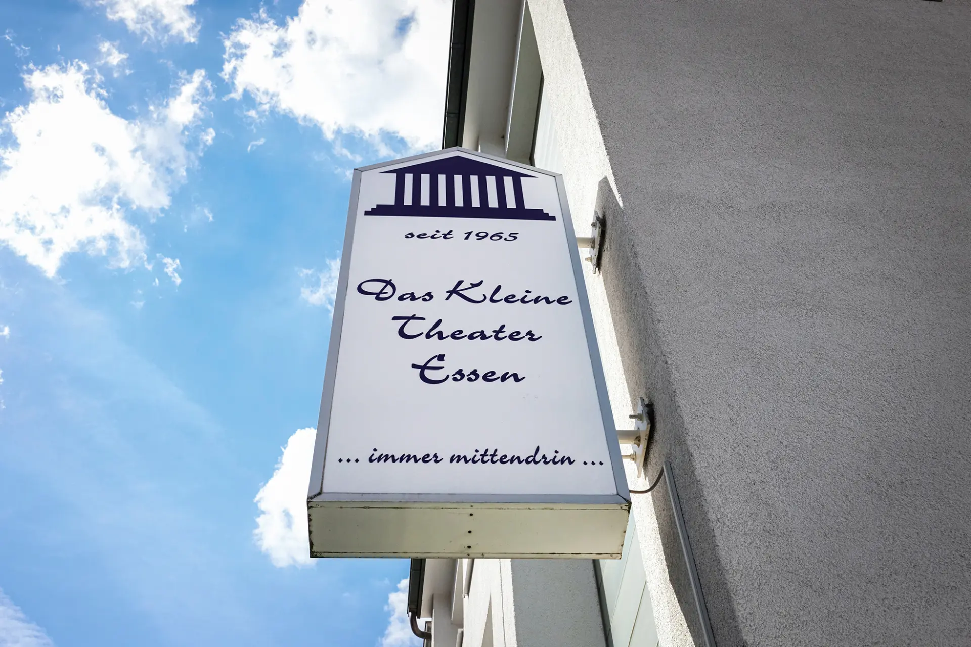 Rechteckiges Schild vom Kleinen Theater Essen vor blauem Himmel, weiß mit lila Streifen.