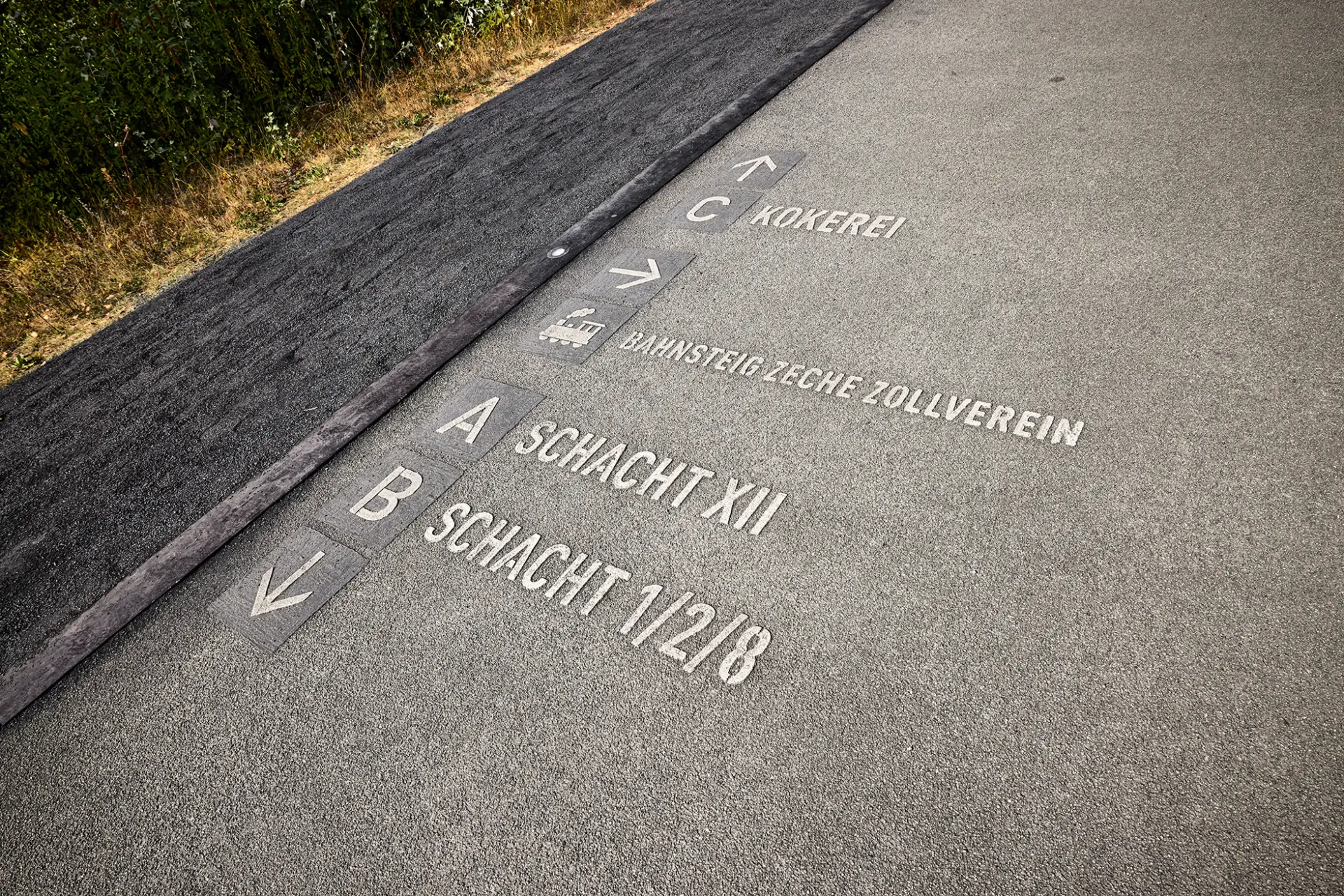 Ein grauer Fußweg mit eingeprägten Wegweisern zur Kokerei und den Schächten XII und 1/2/8 der Zeche Zollverein.