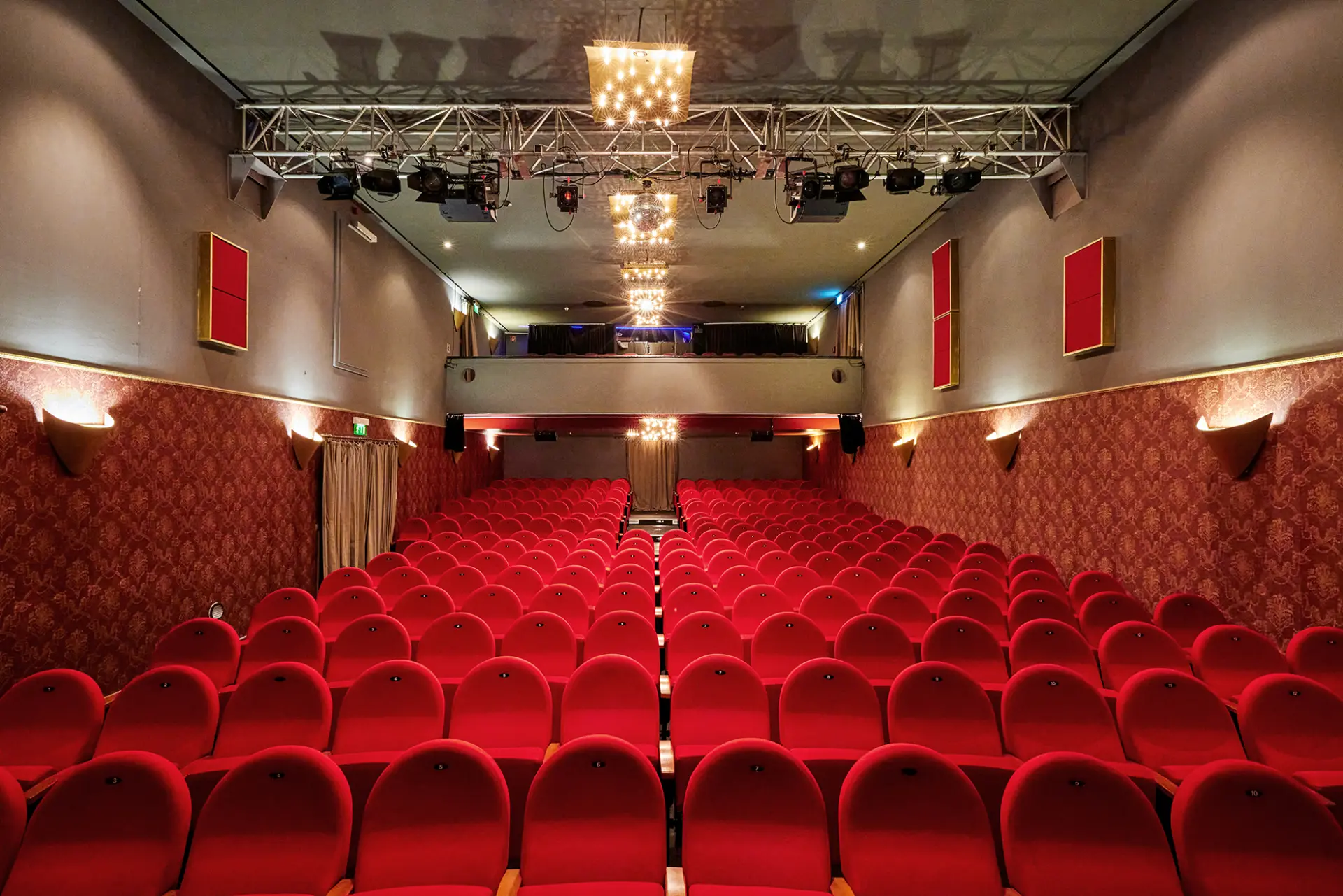 Das Bild zeigt den leeren Zuschauerraum eines Theaters mit roten Polstersitzen und einer Bühne im Hintergrund.