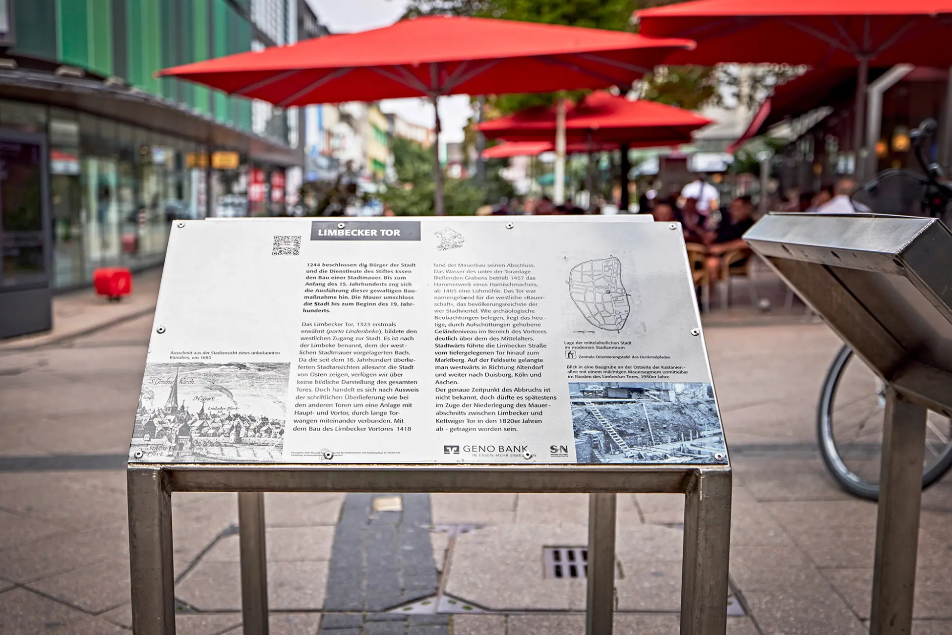 Informationsschild über das Limbecker Tor in Essen mit roten Sonnenschirmen im Hintergrund.