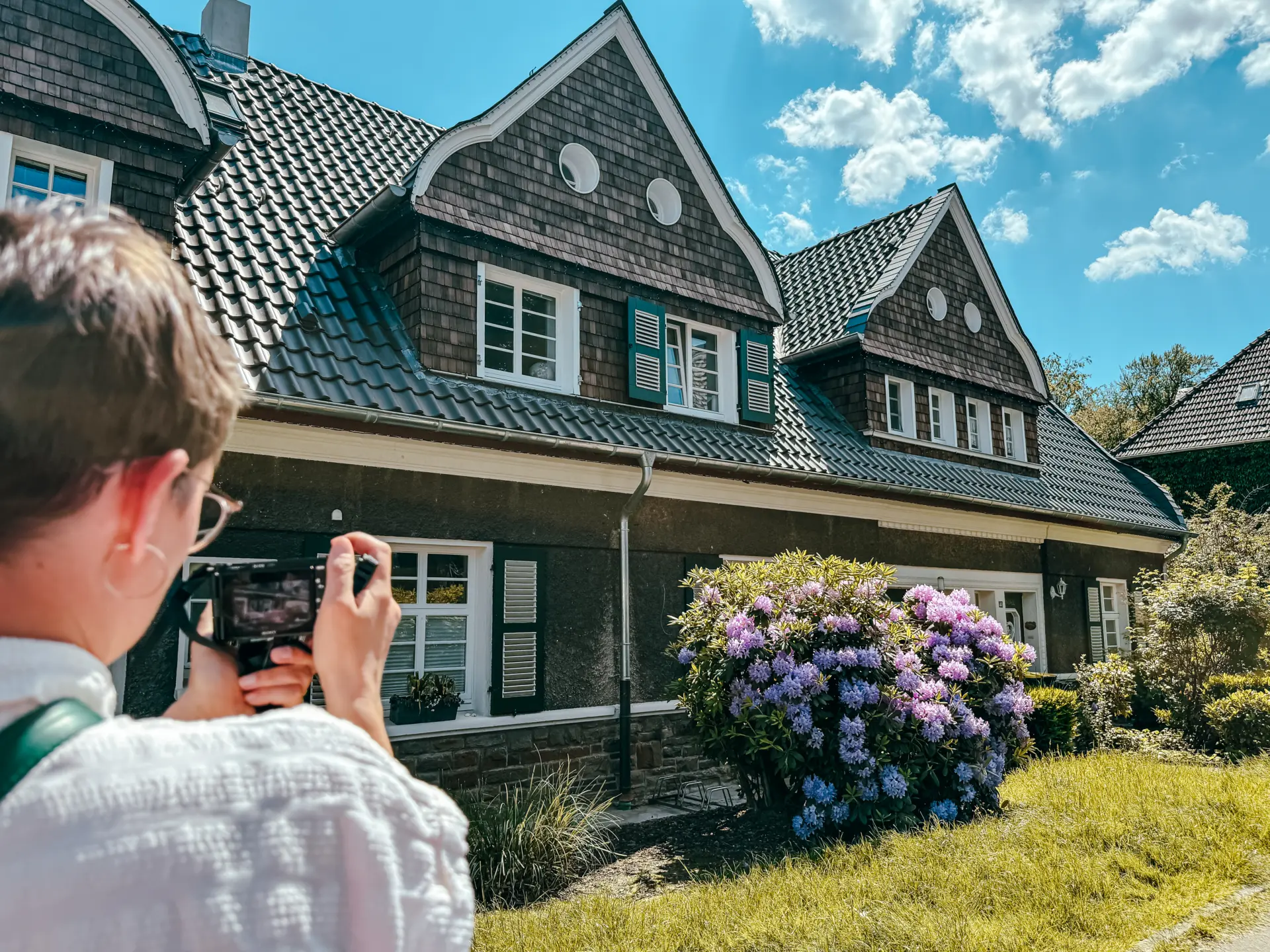 Haus mit Erker, Fotografin davor, blühender Rhododendron und blauer Himmel im Hintergrund.