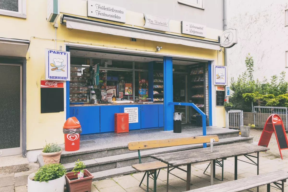 Kiosk mit blauer Fassade, Verkaufsregal und Werbeplakaten, davor Holzbänke und Mülleimer.