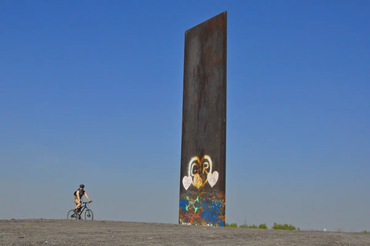 Ein Radfahrer vor einer großen, rostigen Stahlskulptur auf Kies mit blauem Himmel im Hintergrund.