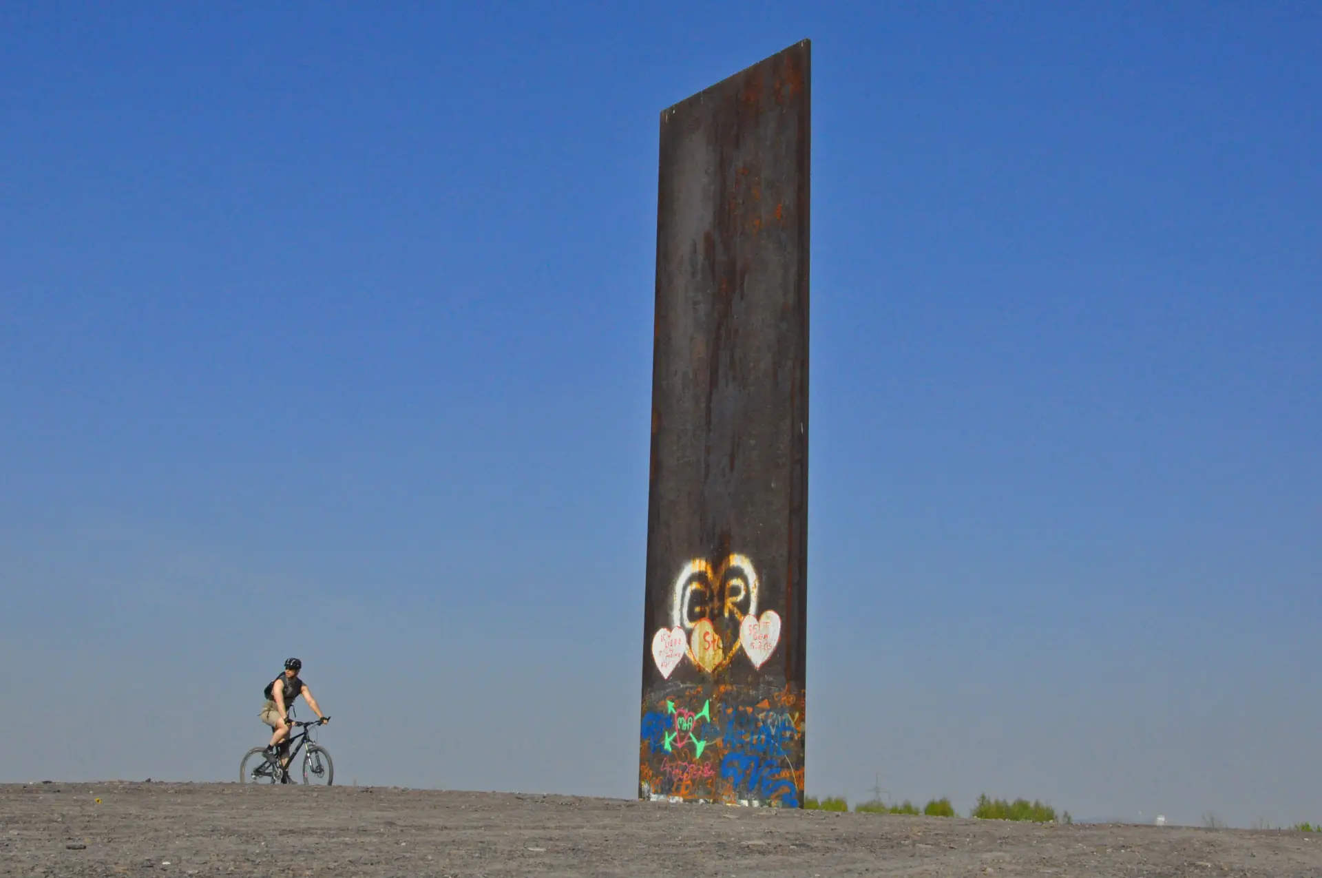 Ein Radfahrer vor einer großen, rostigen Stahlskulptur auf Kies mit blauem Himmel im Hintergrund.