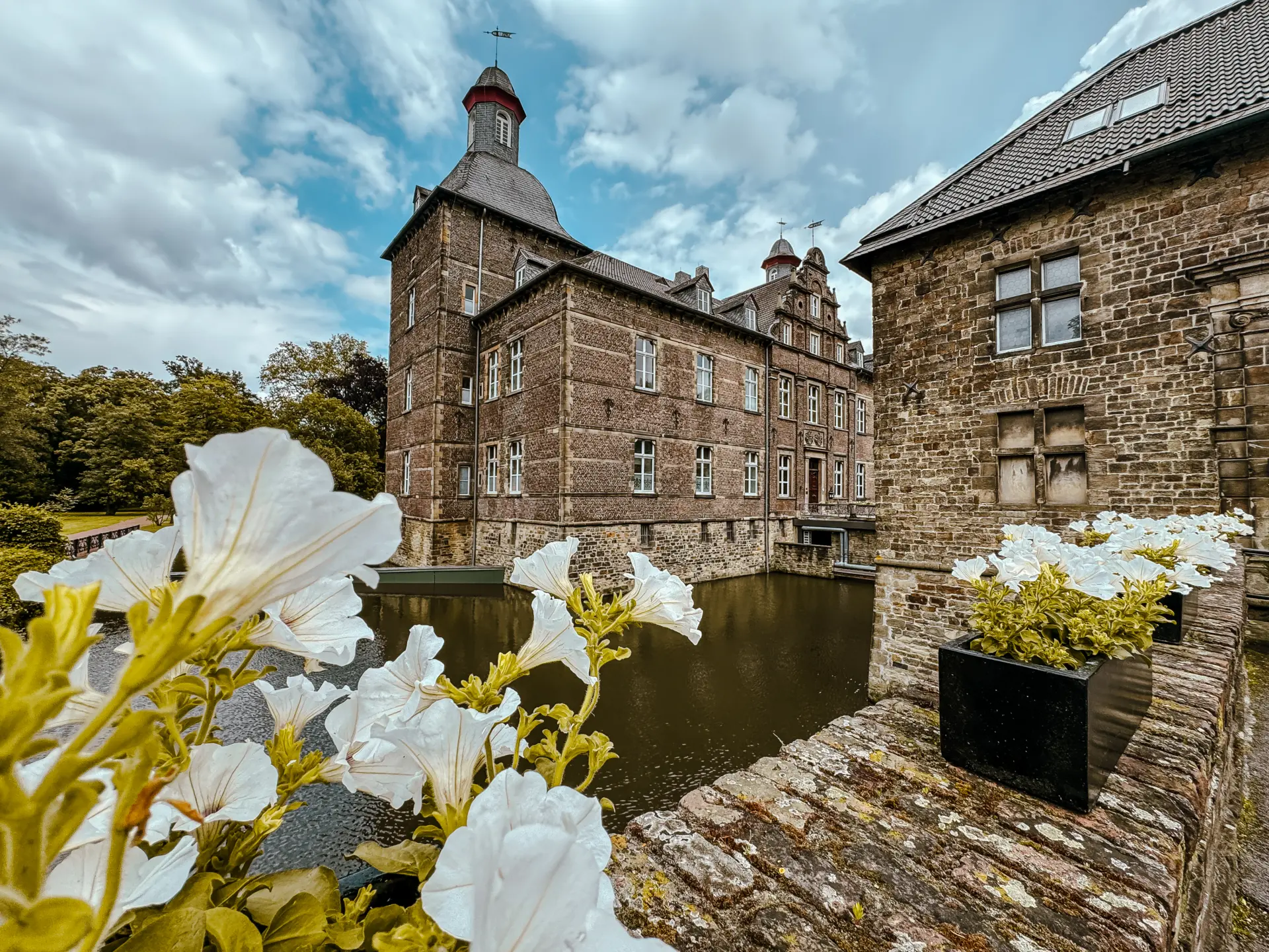 Historisches Backsteinschloss umgeben von Wassergraben und blühenden weißen Petunien im Vordergrund.