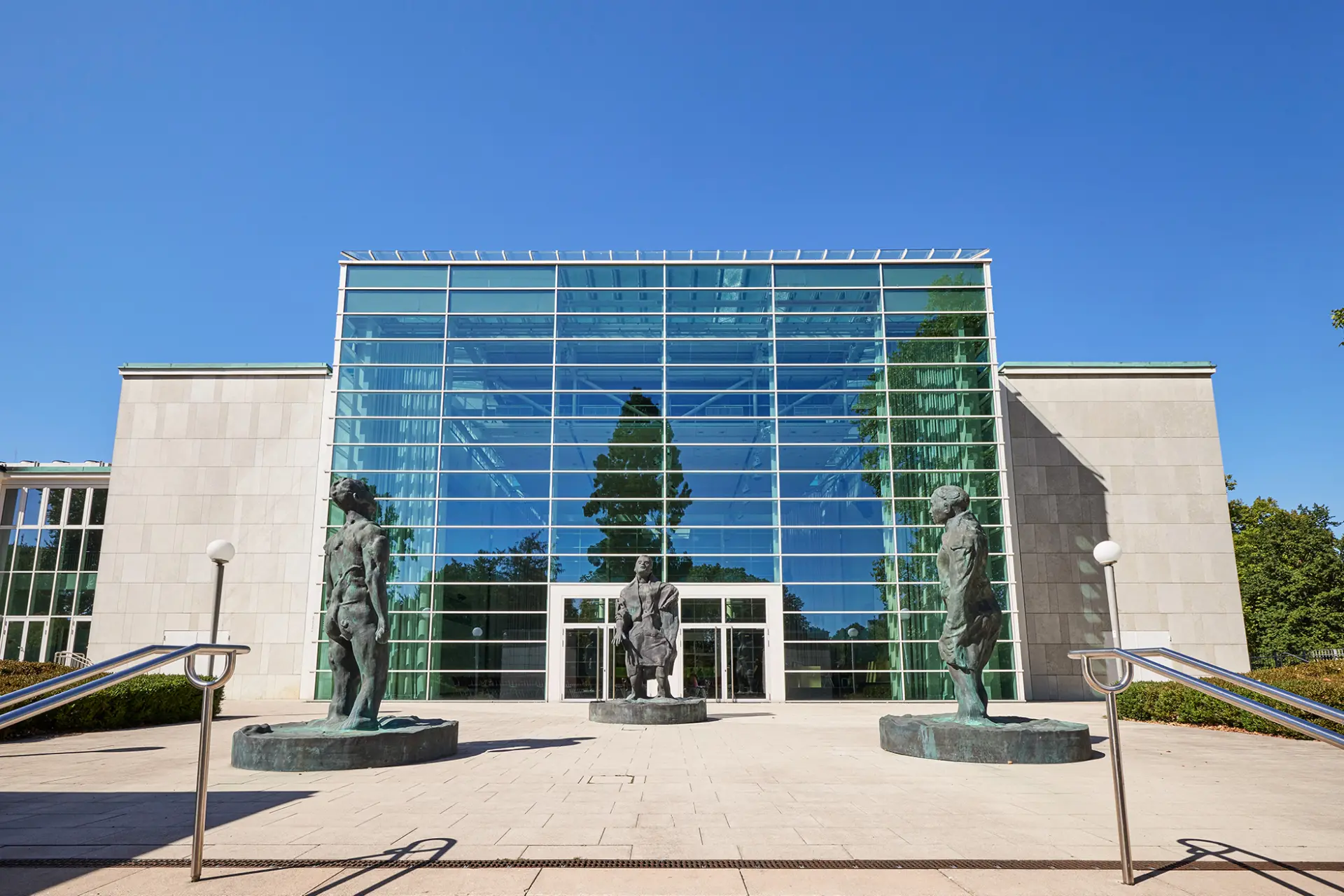 Transparentes Glasgebäude mit Skulpturen im Vordergrund, umgeben von Grünflächen unter blauem Himmel.