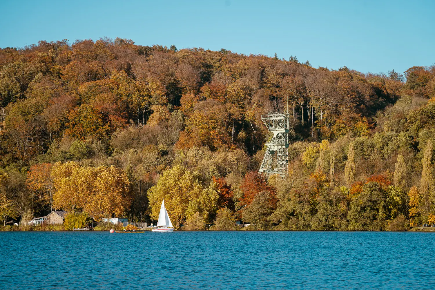 Einsamer Segelboot im ruhigen See vor herbstlich bewaldetem Hügel mit kleinem Gebäudekomplex.