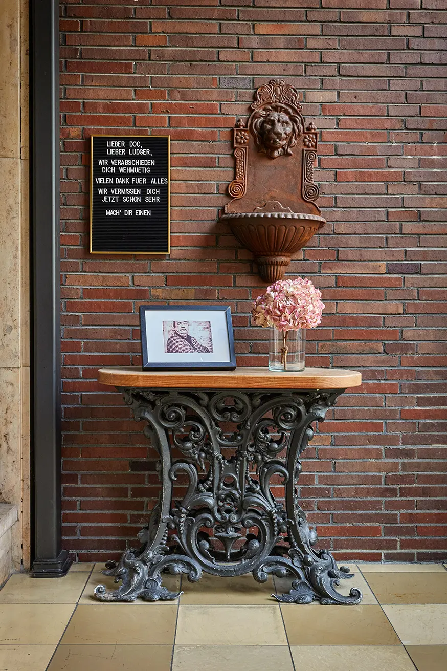Ein antiker Metalltisch mit gerahmtem Bild und Vase steht vor einer Wand mit Ziegeldesign und Relieflöwenkopf.