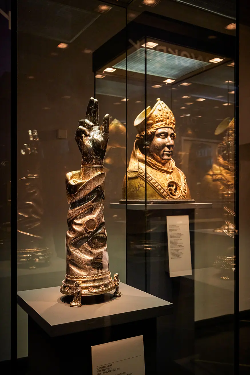 Skulptur einer segnenden Hand aus Silber; im Hintergrund ein vergoldeter Bischofskopf in Glasvitrine.