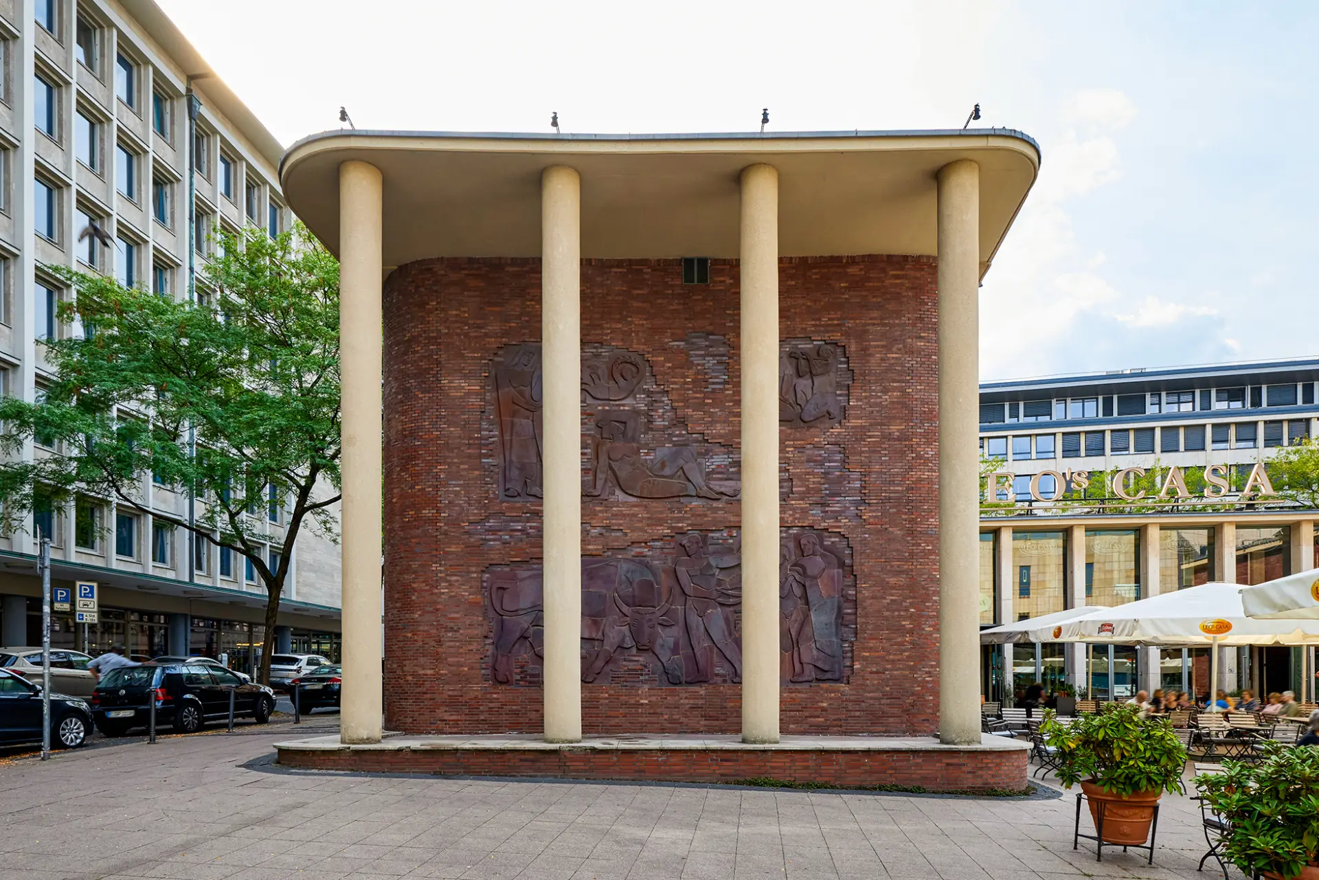 Das Bild zeigt eine Wand mit Reliefs und Säulen, die Teil eines Gebäudes in Essen sind. Vor der Wand sind Pflanzkübel und im Hintergrund stehen zahlreiche Fensterreihen eines weiteren Gebäudes.