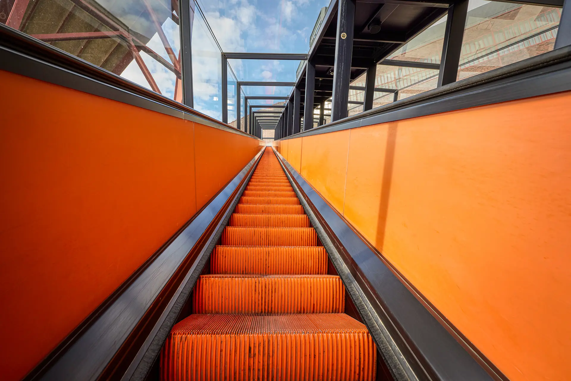 Rolltreppe mit orangen Stufen, eingefasst in schwarze Geländer, führt zwischen gläsernen Wänden ins Freie.