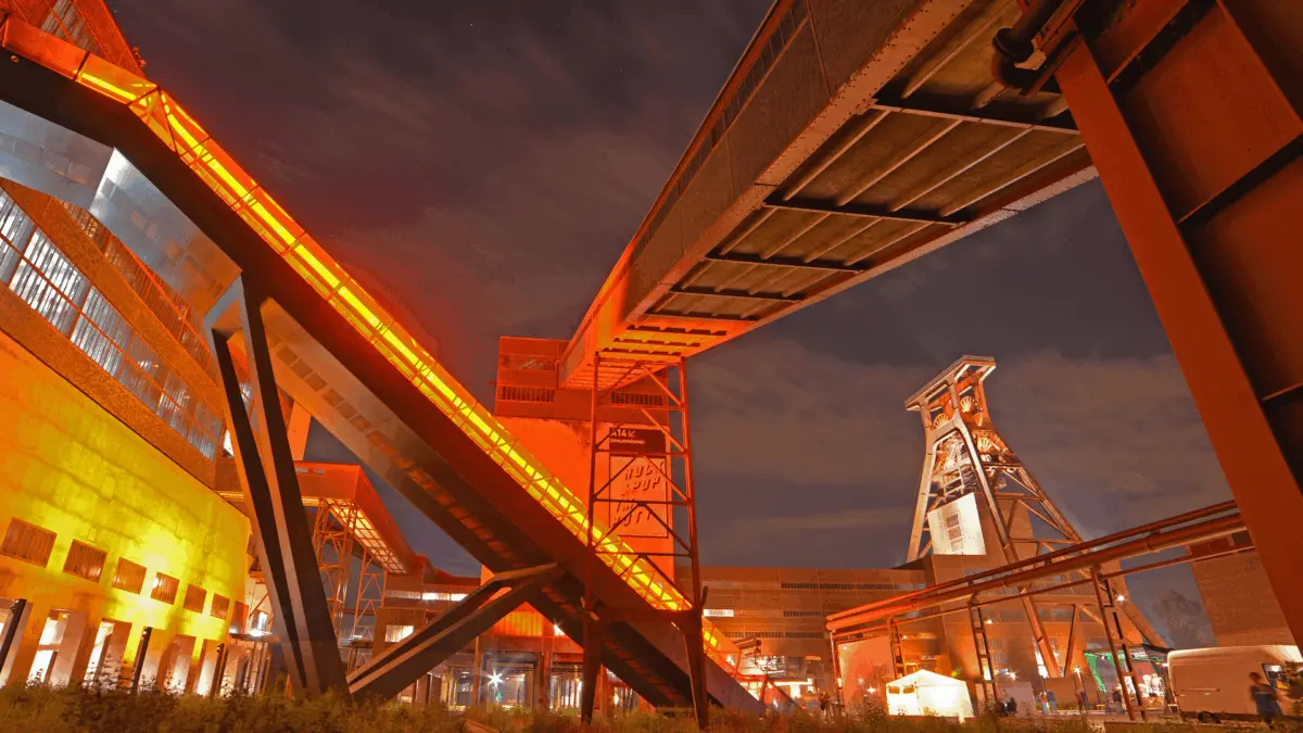 Abendliches Industrieareal mit beleuchteter Förderanlage und markanten Stahlstrukturen.