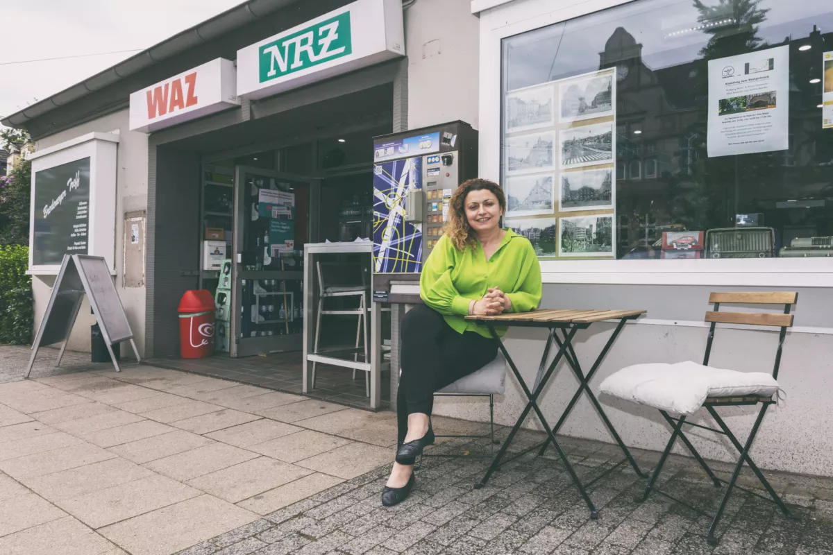 Frau in grüner Bluse sitzt vor einem Zeitungskiosk mit "WAZ" und "NRZ" Schildern an einem Tisch.