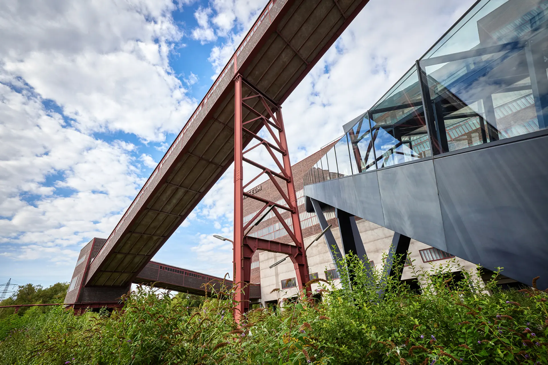 Industriedenkmal mit modernem Glasbau und roten Stahlträgern, blauer Himmel und grüne Vegetation.