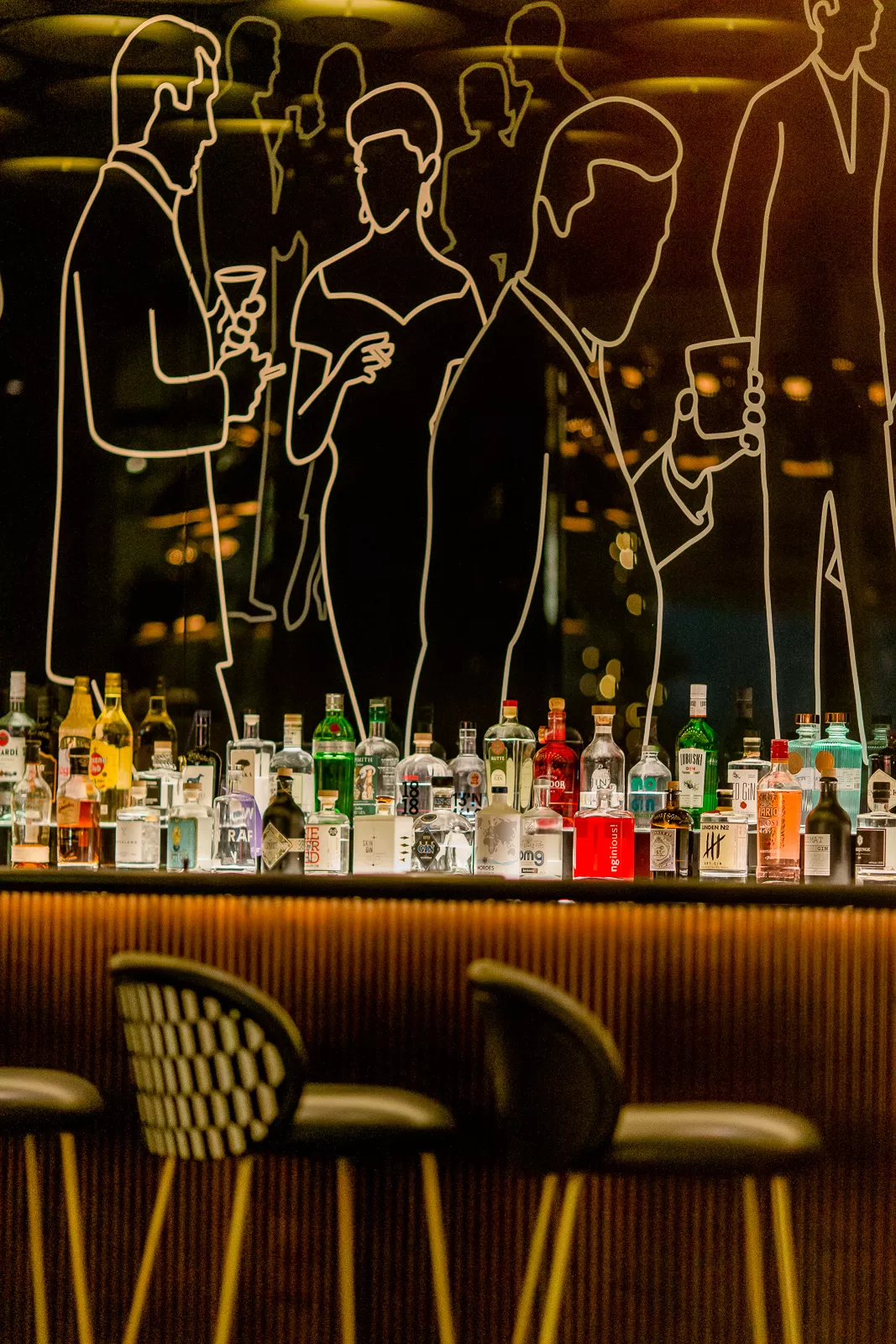 Bar mit eleganten Linienzeichnungen an der Wand, beleuchtete Flaschen und schlichte Barhocker im Vordergrund.