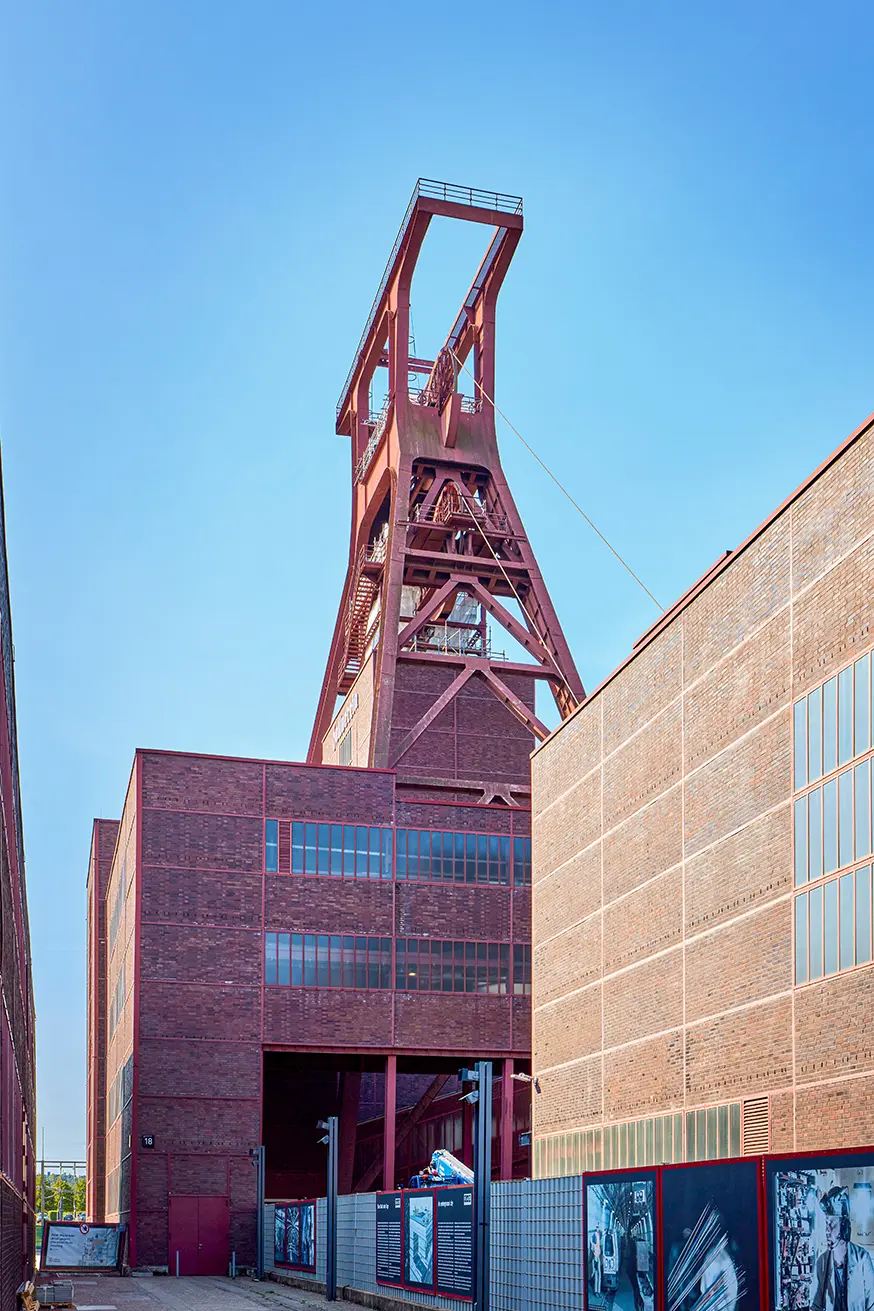 Roter Förderturm der Zeche Zollverein in Essen, umgeben von Backsteingebäuden unter blauem Himmel.