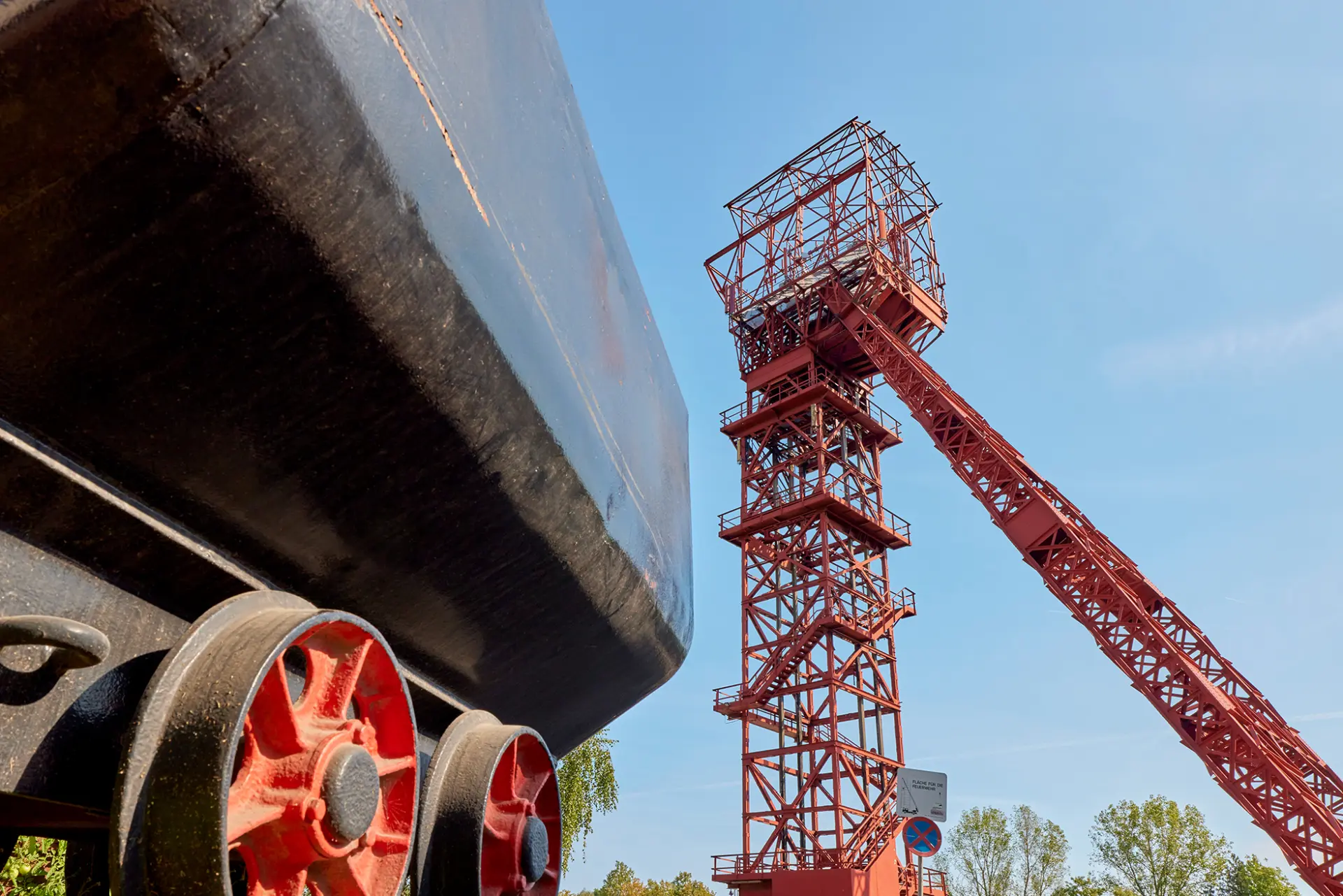 ```html Roter Förderturm und Kohlenwagen vor blauem Himmel, symbolisiert Bergbaugeschichte in Essen. ```