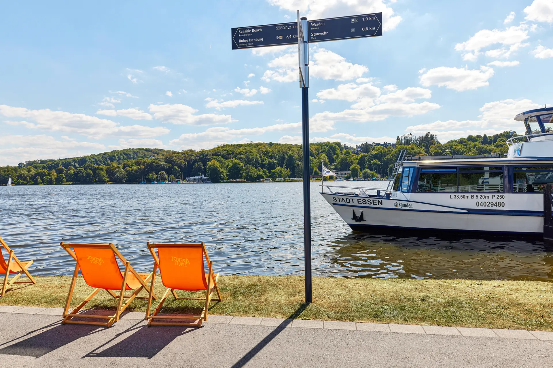 Stadt Essen: Drei orange Liegestühle am Baldeneysee mit einem Ausflugsschiff im Hintergrund.