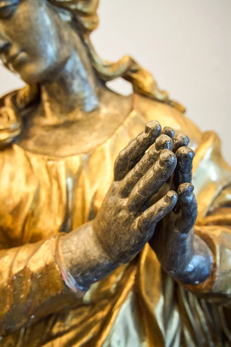 Goldfarbene Statue einer betenden Frau in einem ruhigen Ausdruck, Nahaufnahme der gefalteten Hände.