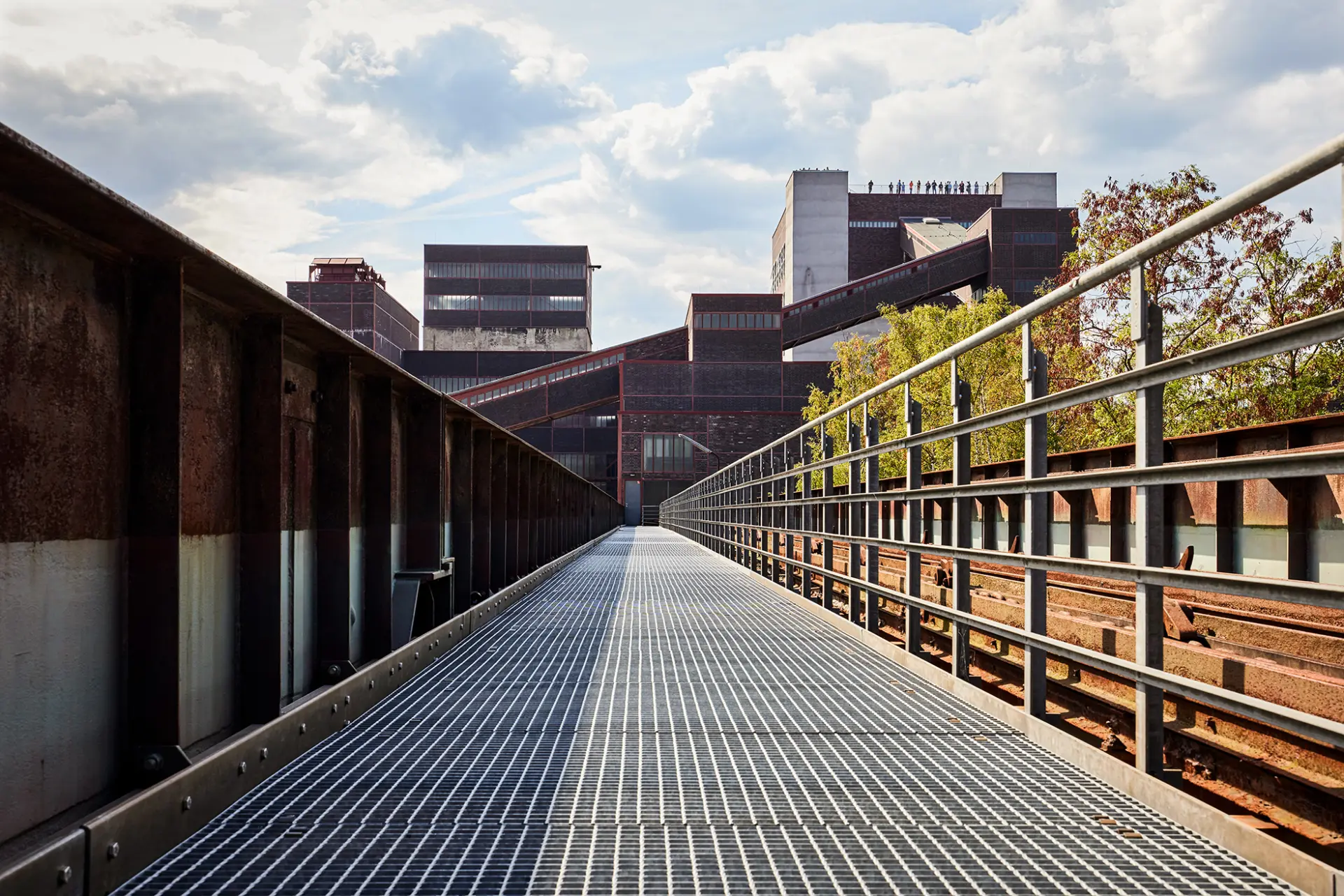 Zollverein-Kokerei: Stahlsteg führt zu Industriegebäuden, von Bäumen flankiert, mit wolkigem Himmel.