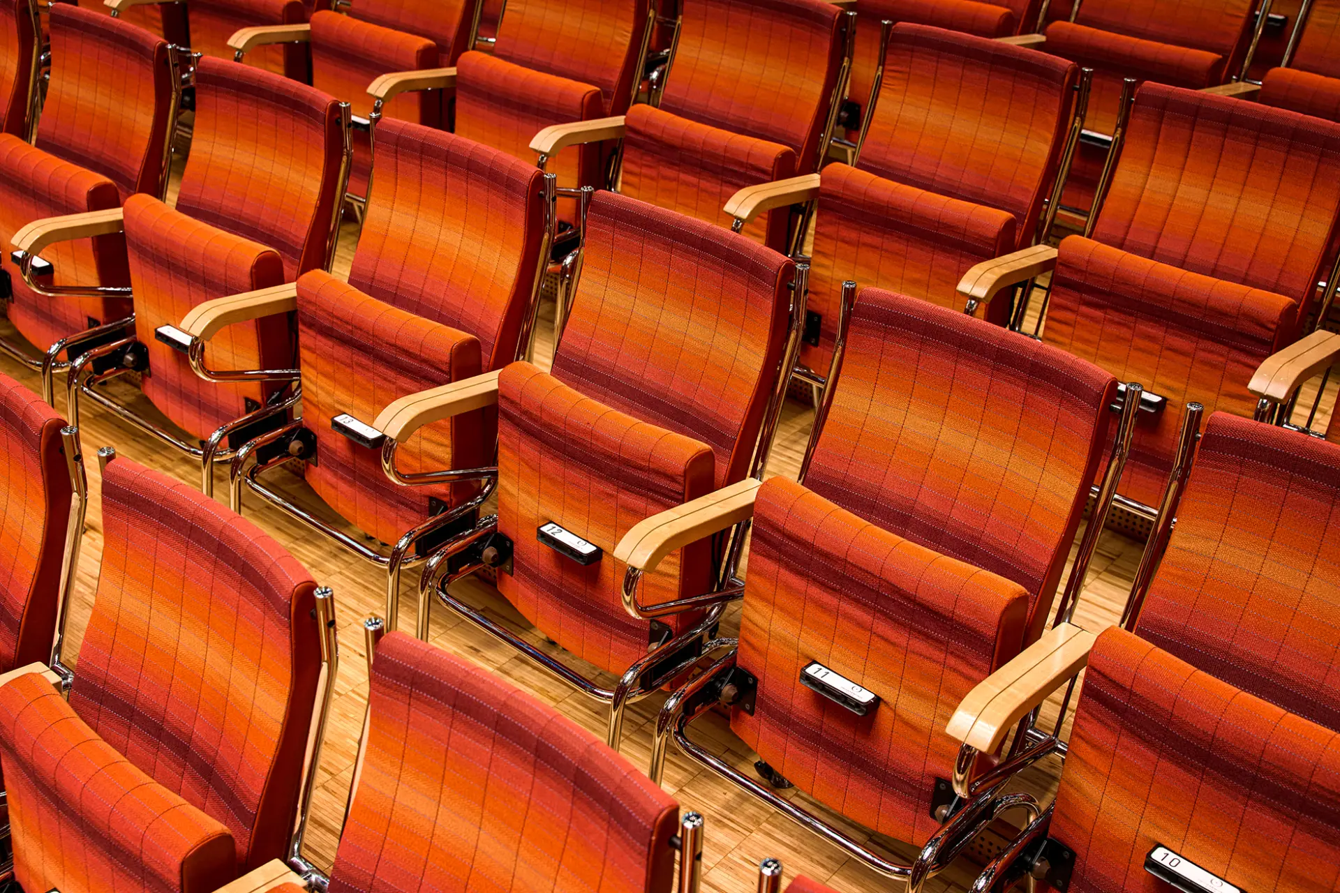 Reihen von leeren, orange-rot gepolsterten Stühlen in einem Konzertsaal, mit Holzboden.