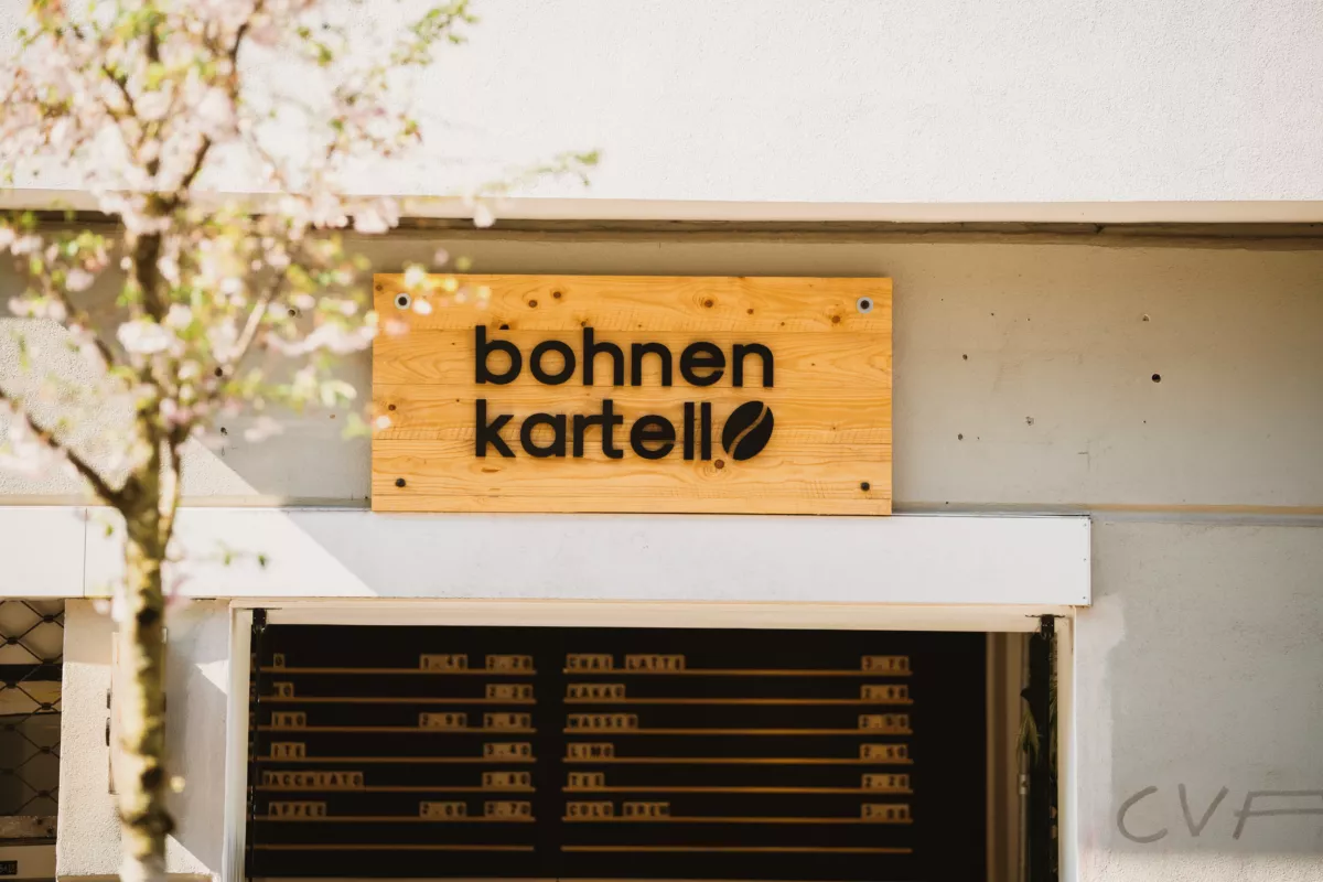 Holzschild "Bohnen Kartell" über einer modernen Fassade, Baum mit zarten Blüten im Vordergrund.
