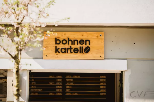 Holzschild "Bohnen Kartell" über einer modernen Fassade, Baum mit zarten Blüten im Vordergrund.