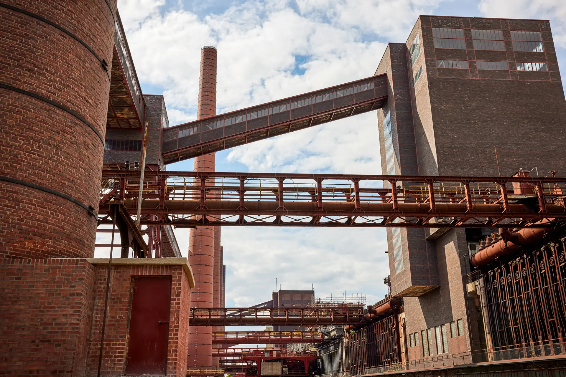 Rote Backsteinindustriegebäude mit Rohren und Laufstegen, wolkiger Himmel im Hintergrund.