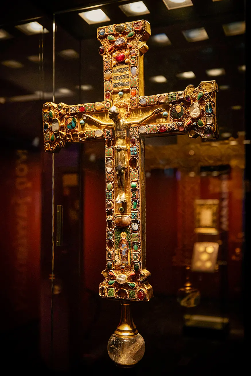 Kunstvoll verziertes, goldenes Kruzifix mit Edelsteinen, präsentiert in einem Museum.