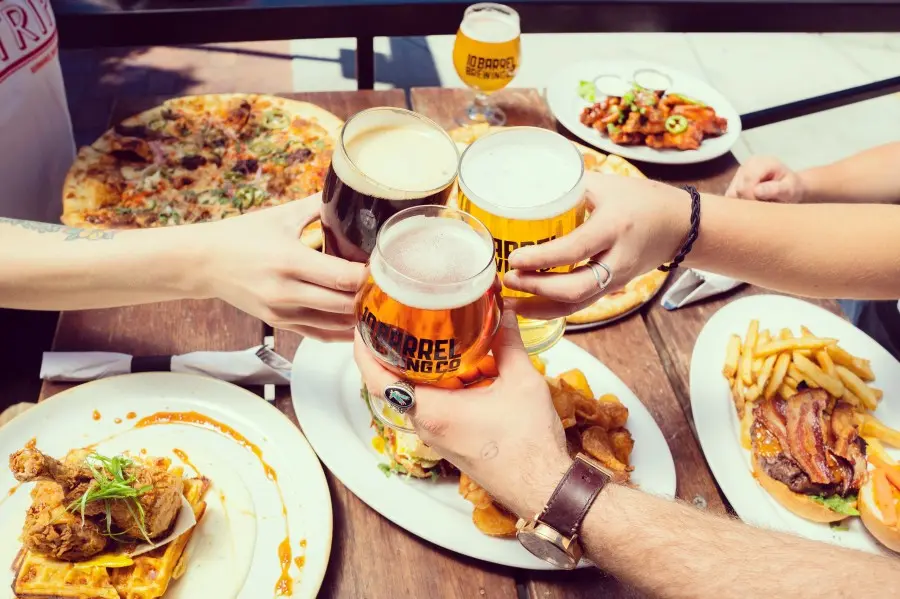 Freunde stoßen mit Bier an, auf dem Tisch Pizza, Burger und Pommes