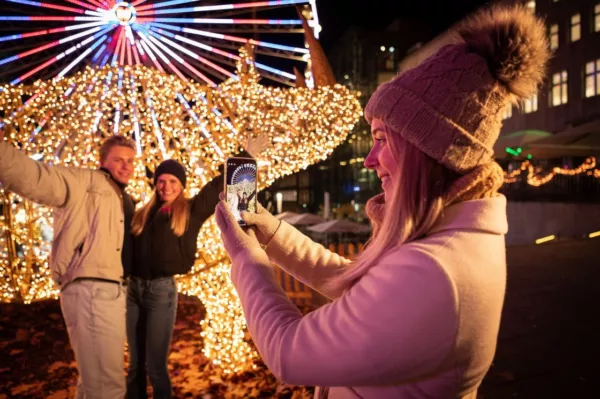 Eine junge Frau mit Pudelmütze fotografiert mit dem Smartphone zwei Personen vor einer leuchtenden Lichterfigur der Essener Lichtwochen, dahinter ein beleuchtetes Riesenrad